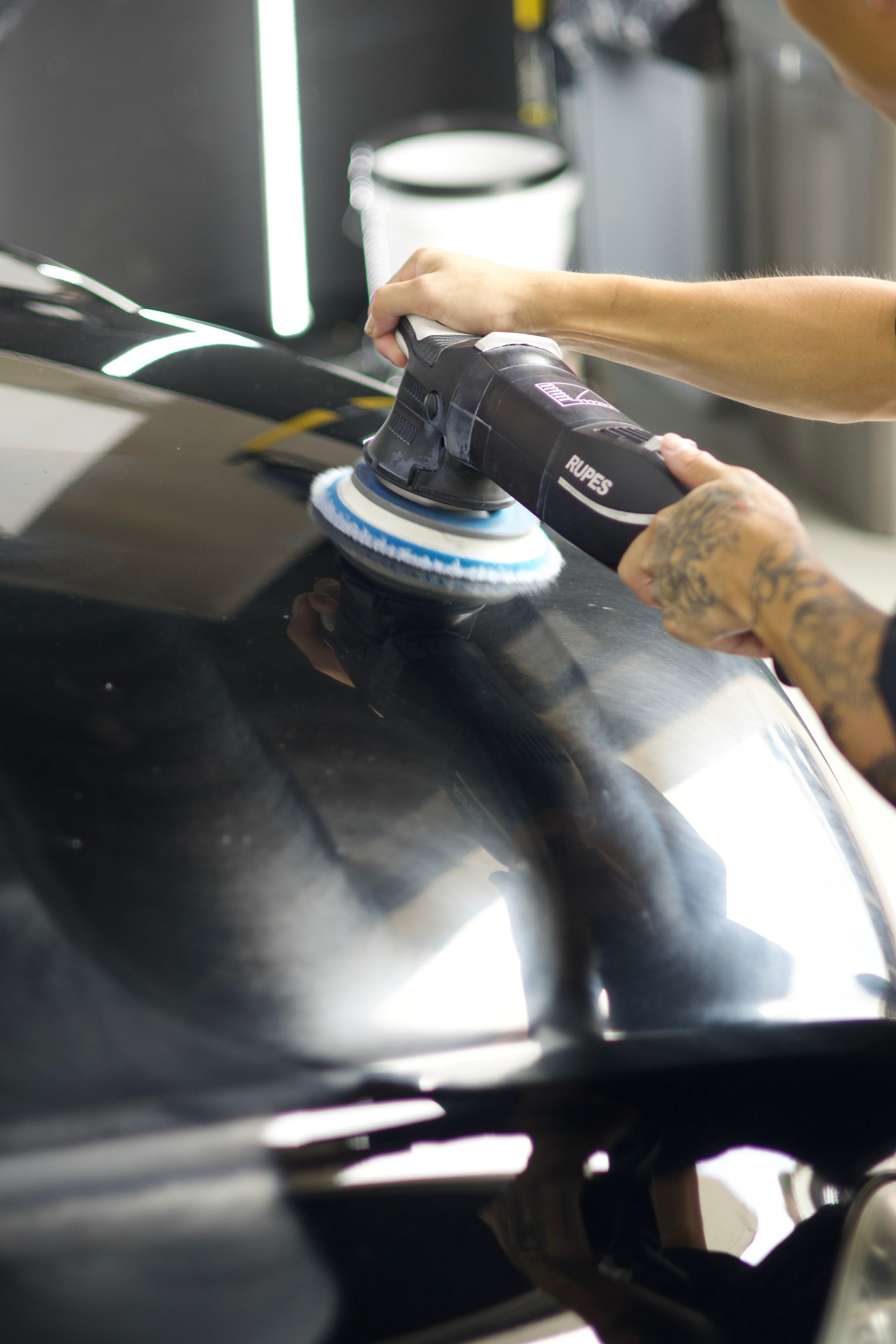 Person polishing a glossy black car hood with a buffer in a workshop