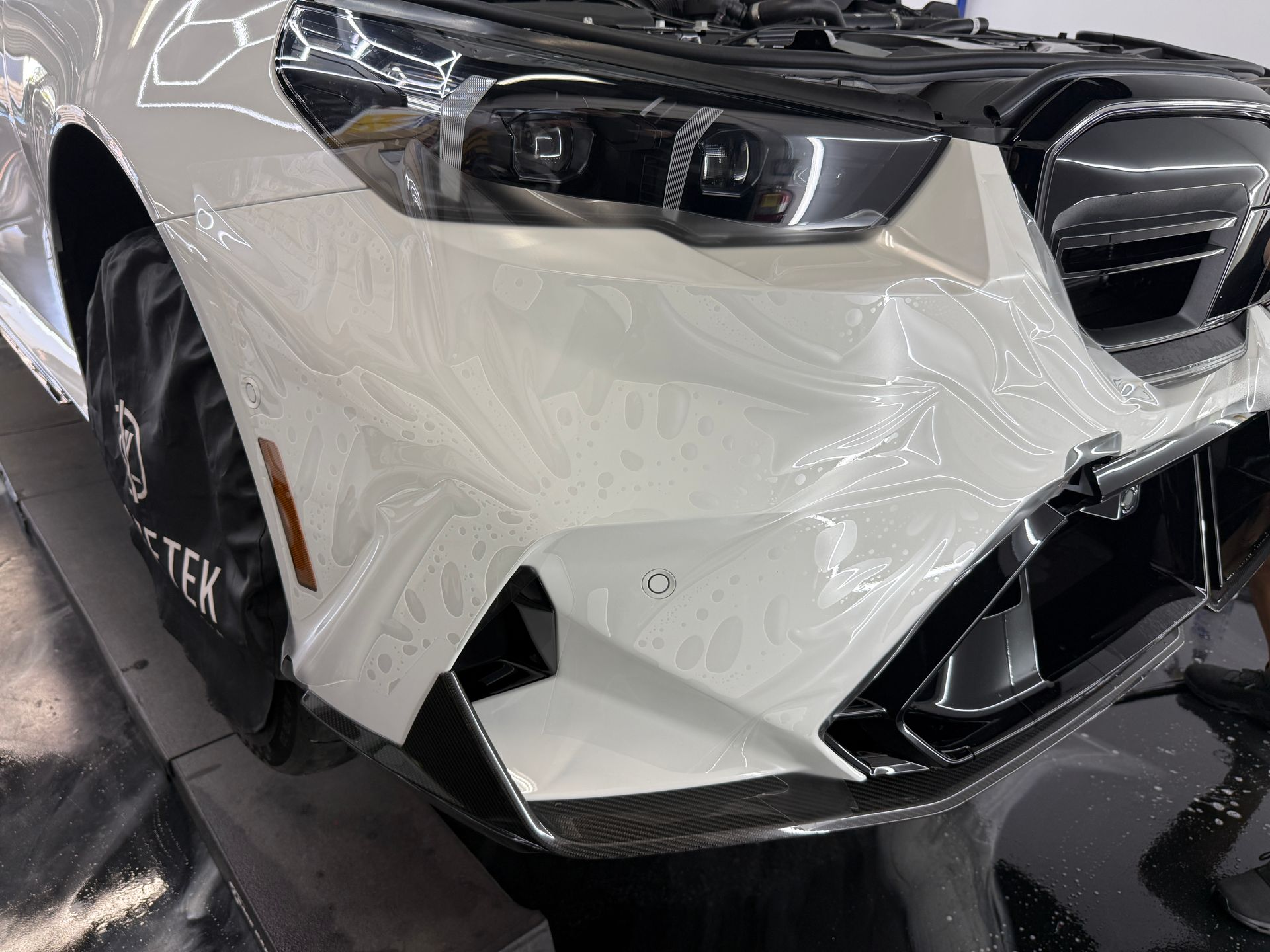 Front-left view of a white BMW with a damaged bumper and crumpled hood at an indoor auto shop