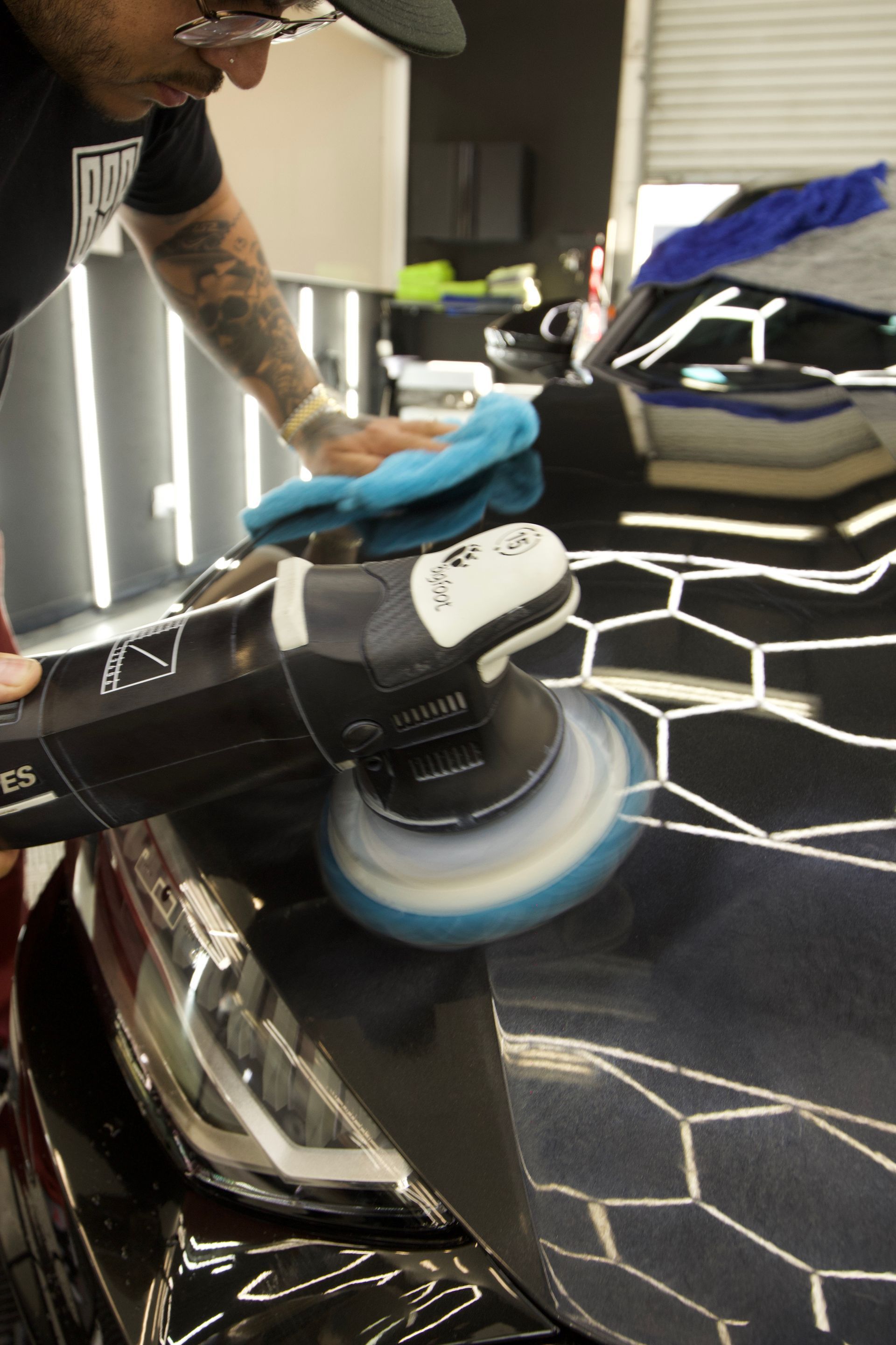 Person polishing a black car hood with a buffer and blue cloth in a garage