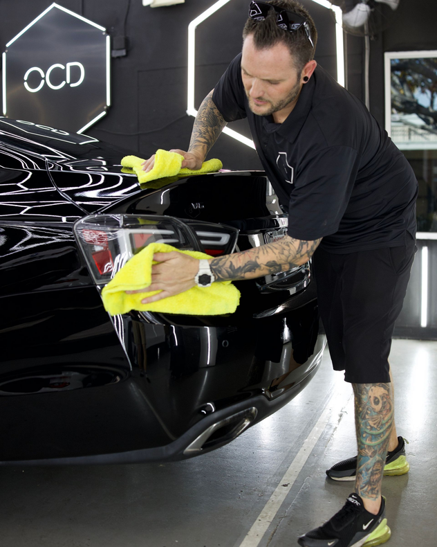 Man cleaning a black Audi with yellow microfiber cloths in a glossy showroom