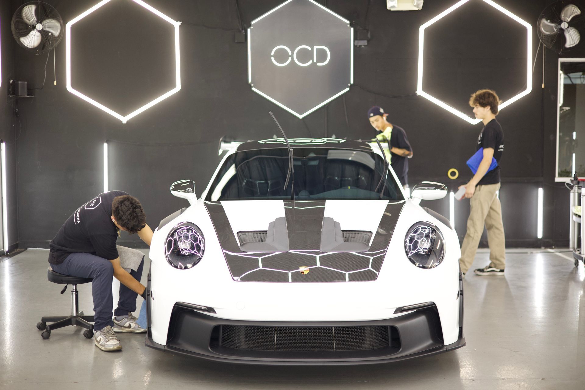 White sports car in a studio with three people working around it and neon hexagon lights on the wall
