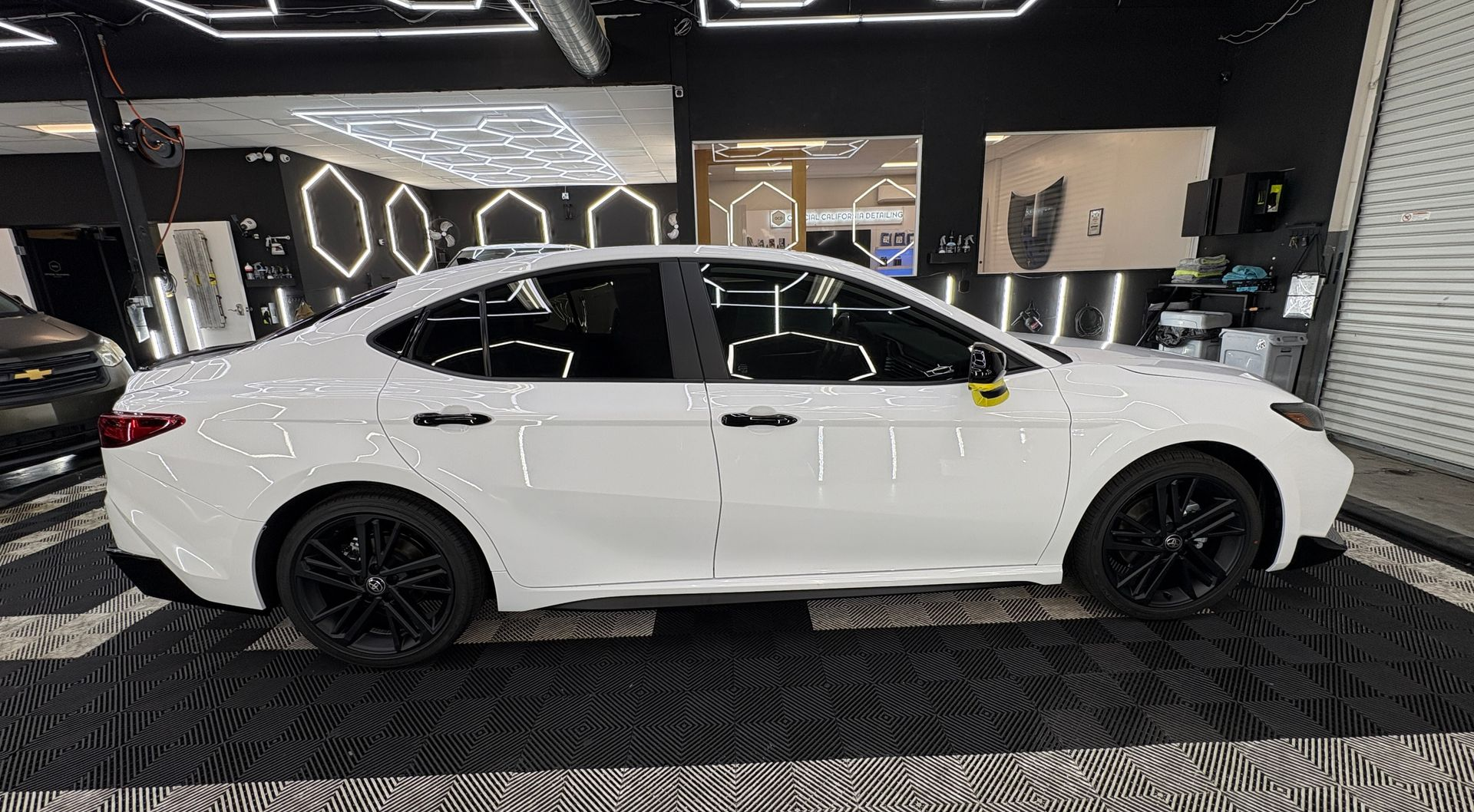 White sedan displayed indoors in a showroom, side view with black wheels and modern lighting.