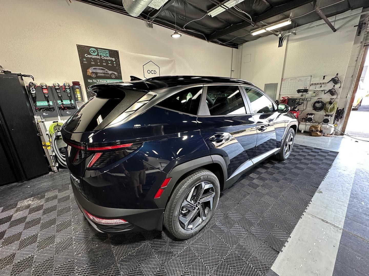 Black SUV in a garage with shop equipment and tire racks, viewed from the rear side.