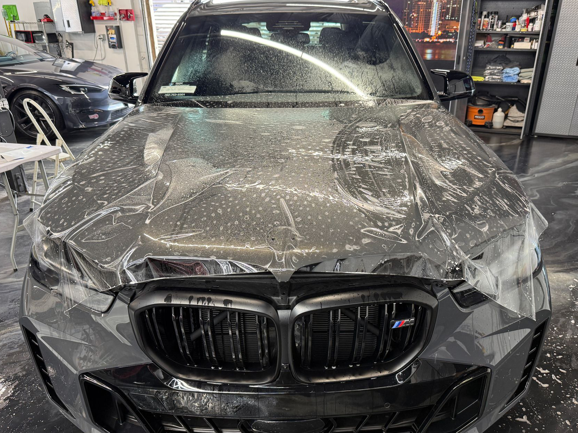 Black BMW in a garage with the hood and front end heavily damaged and covered in dust/spray.