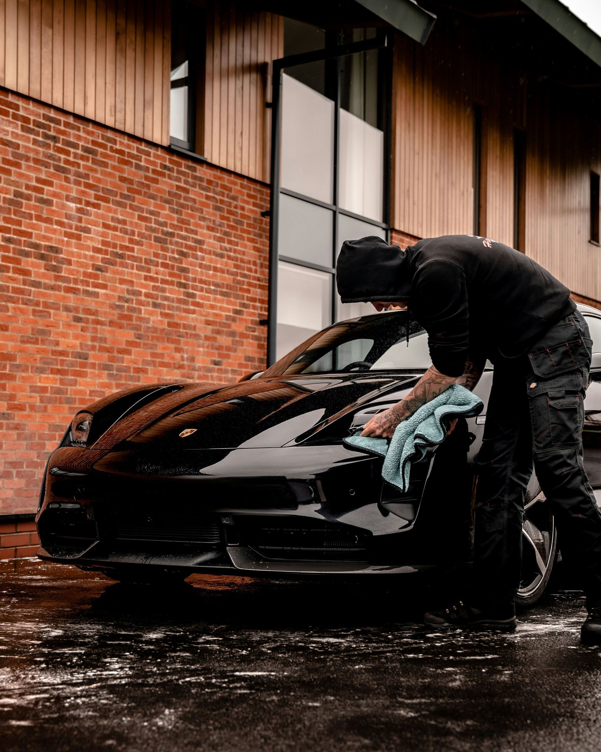 Person washing a black sports car outdoors beside a brick building in the rain