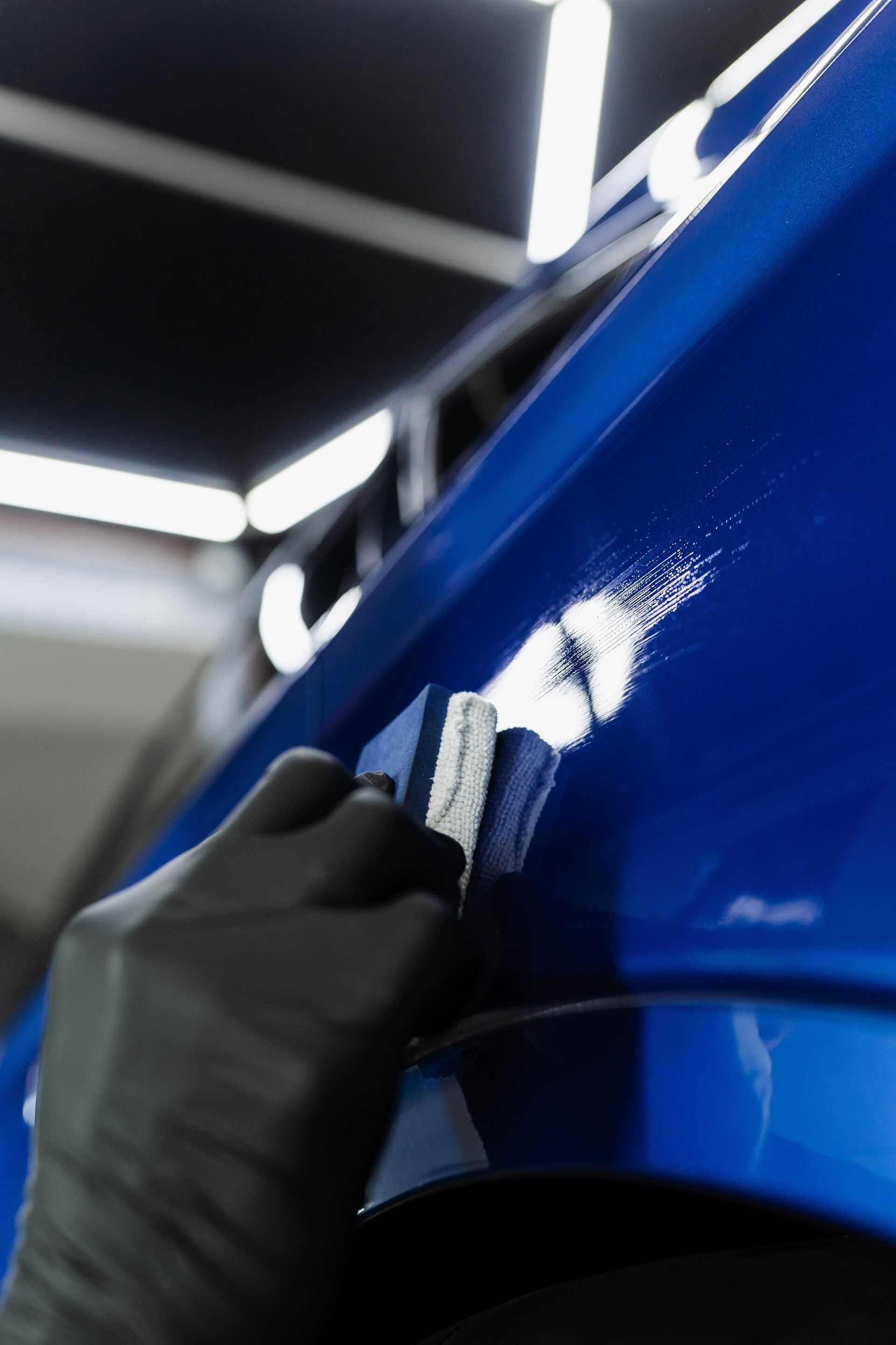 Gloved hand polishing a glossy blue car panel under bright workshop lights