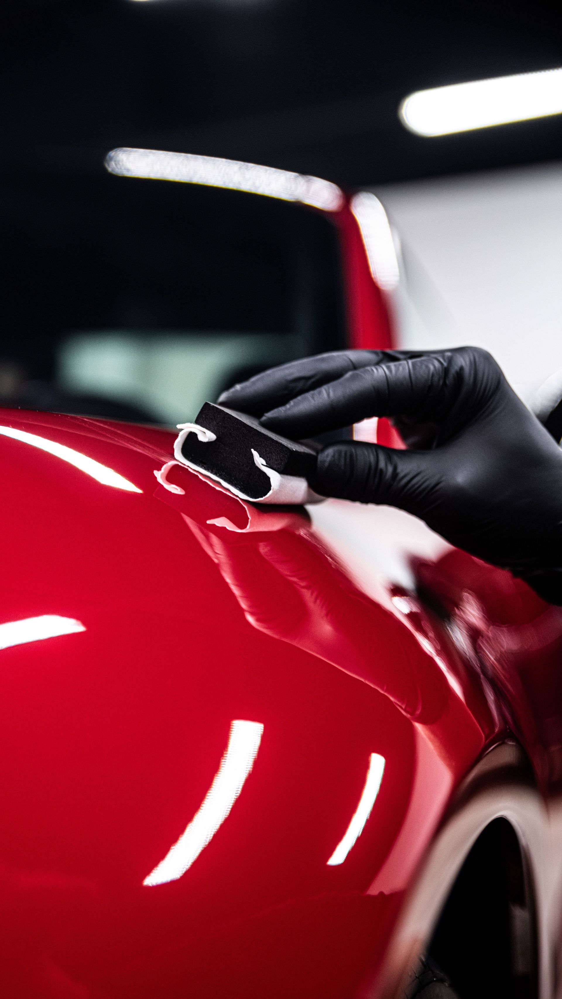 Gloved hand polishing a glossy red car panel under bright workshop lights