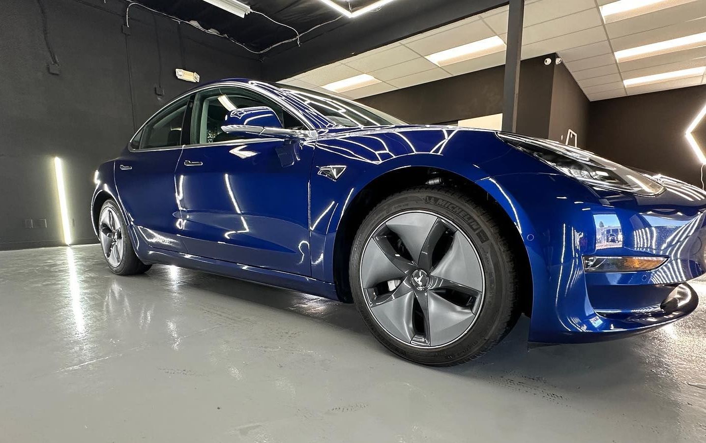 Blue Tesla sedan parked indoors under bright showroom lights, seen from a low angle.