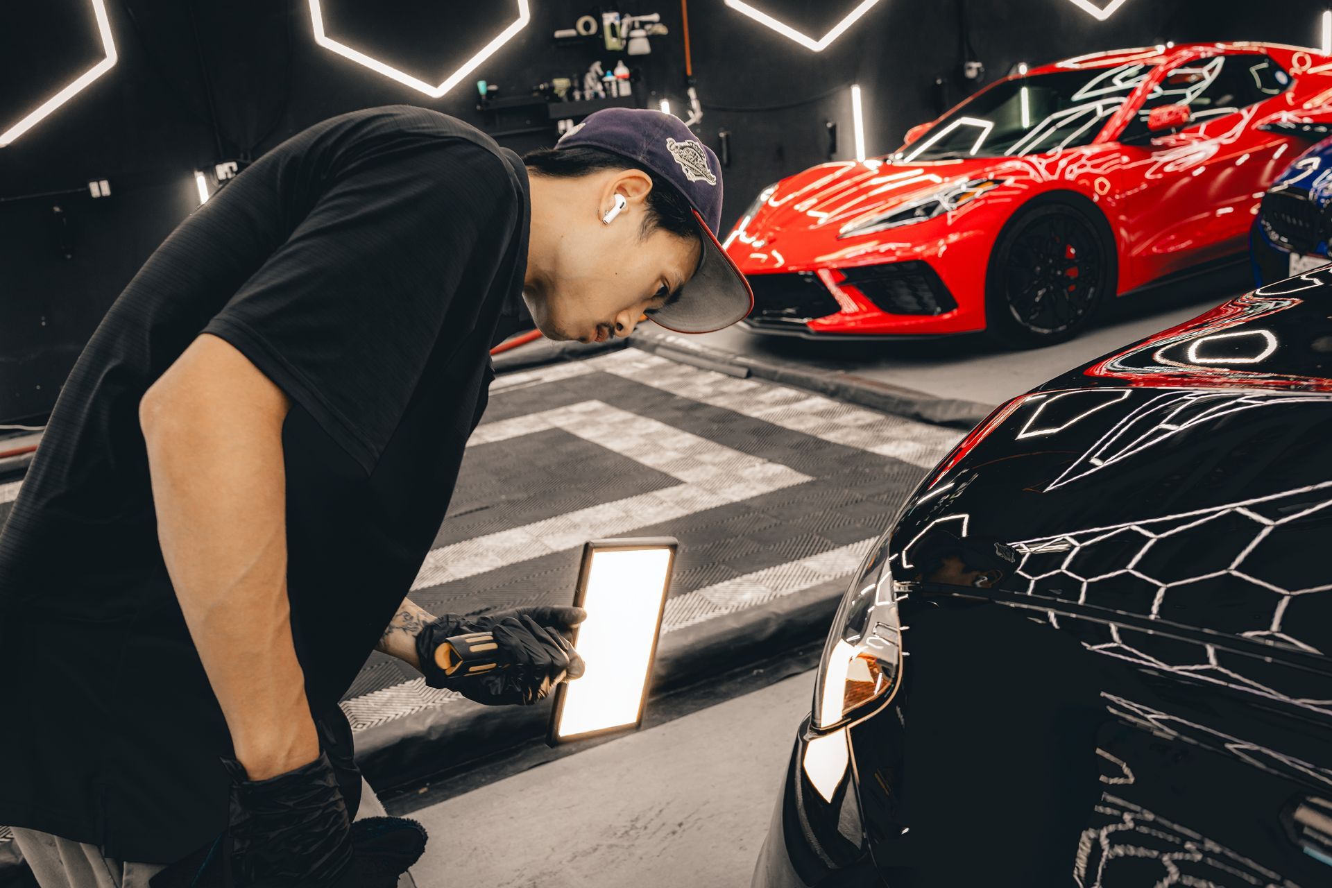 Person inspecting a black sports car under studio lights, with a red sports car in the background