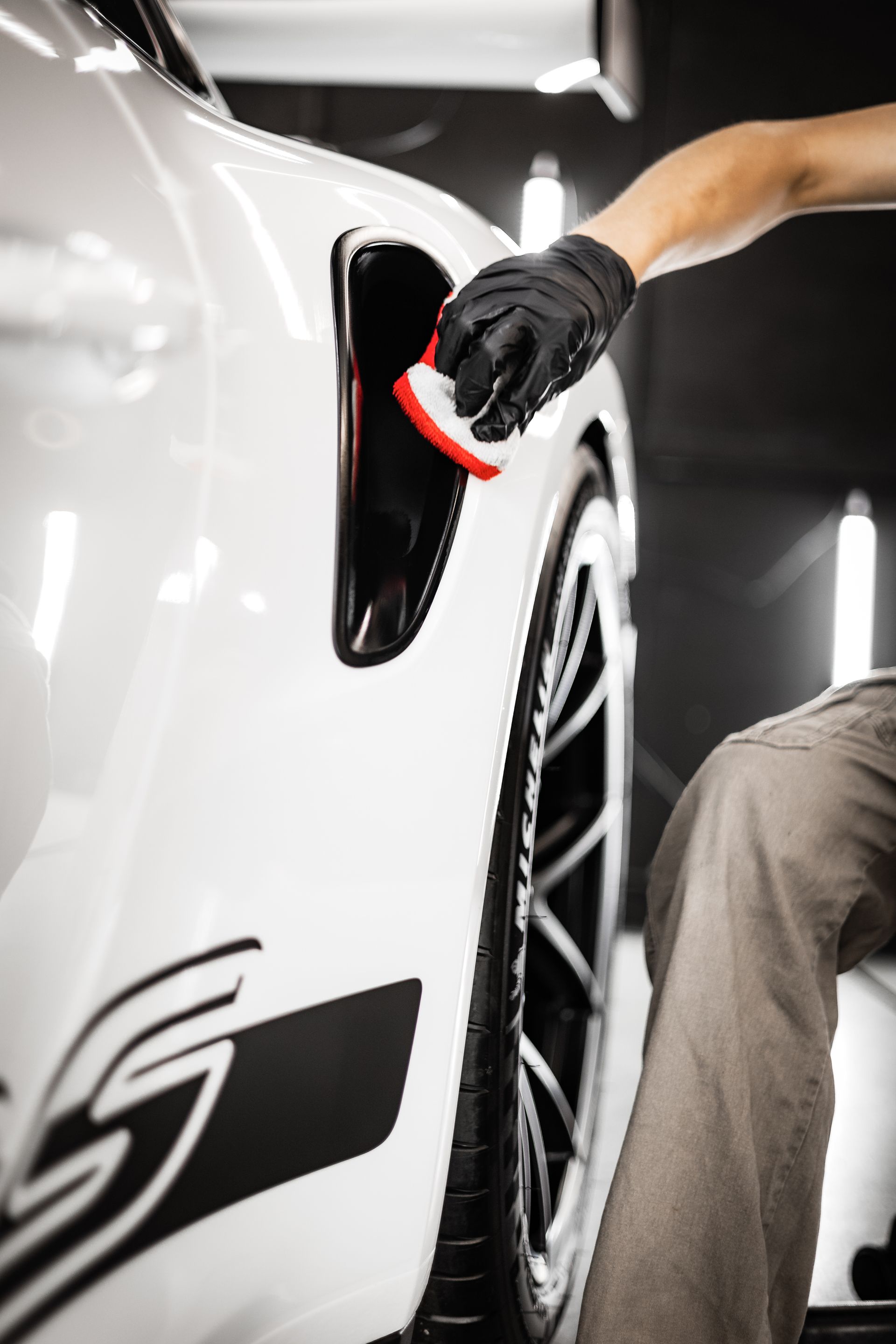 Gloved hand polishing a white sports car’s side panel beside a wheel in a studio.