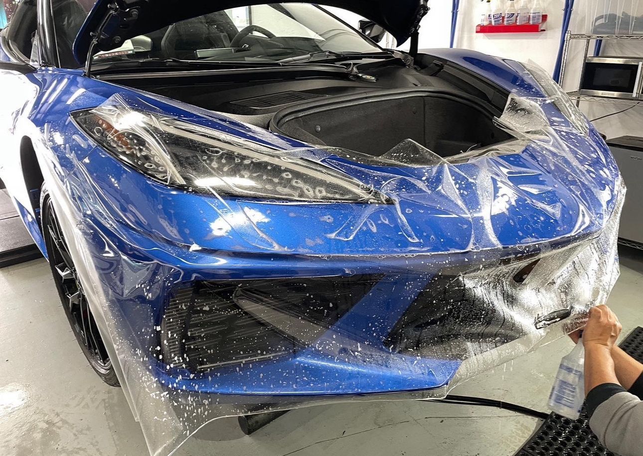 Blue sports car being wrapped in clear protective film in a garage, hood open.