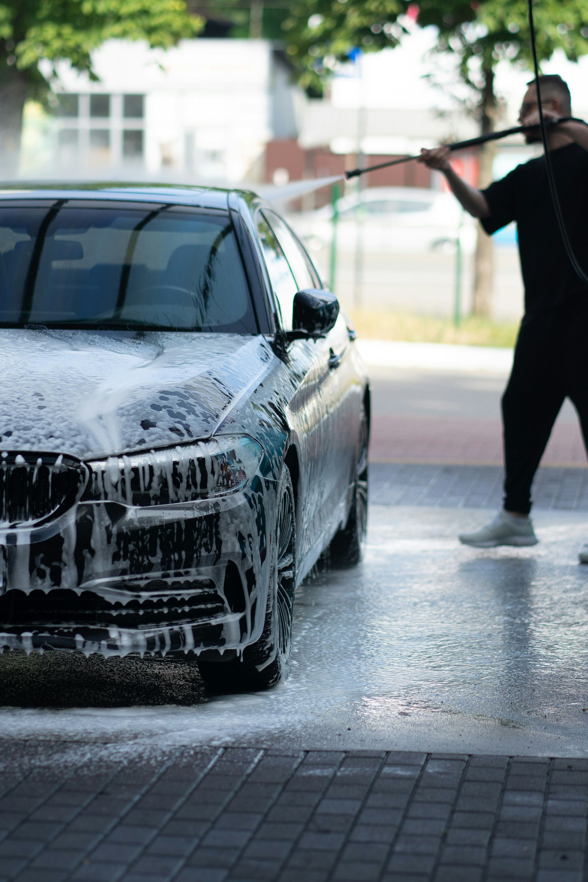 Person washing a black car with a hose in a driveway, covered in soap suds.