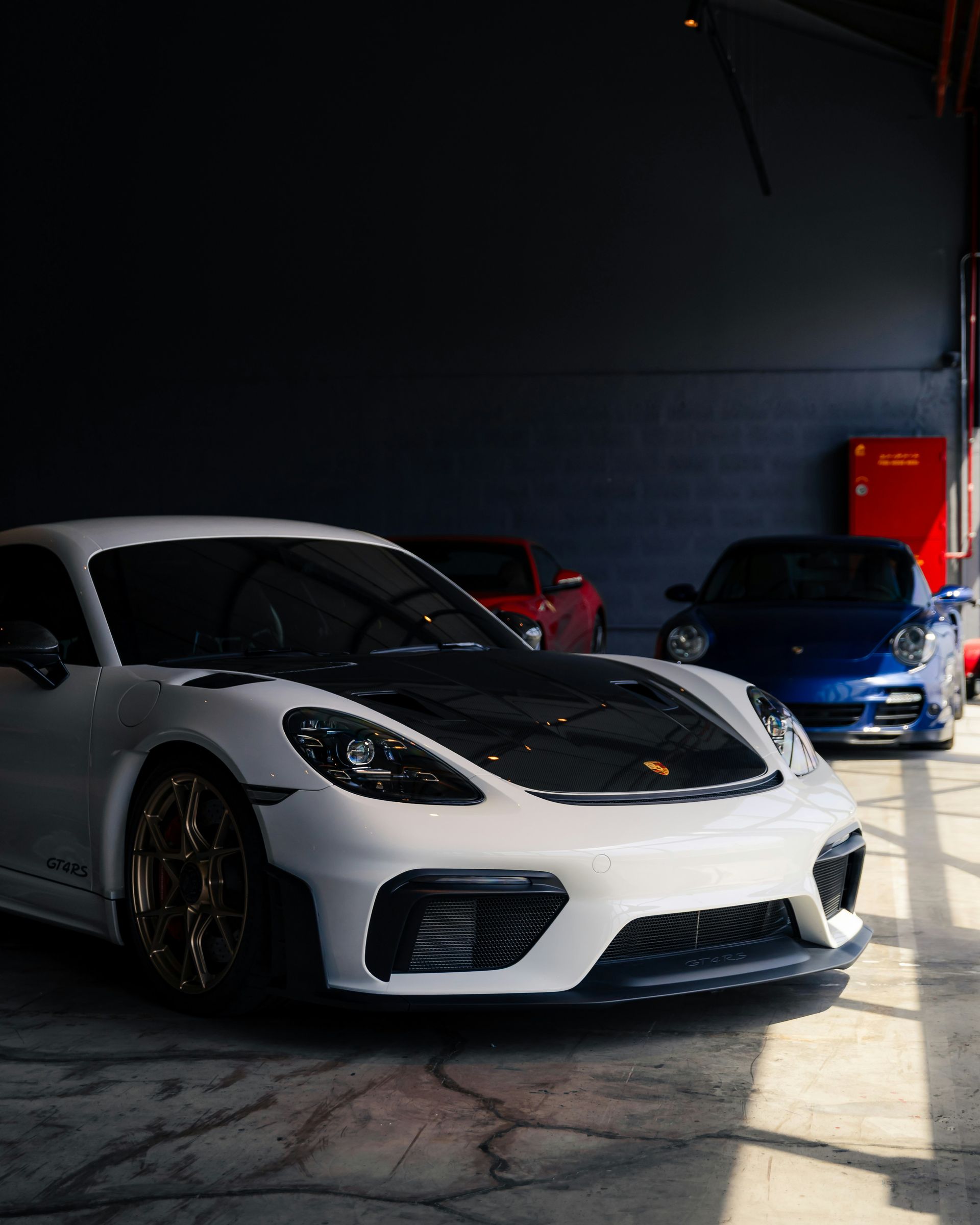 White sports car parked in a dim garage beside a blue car, with bright sunlight on the floor.
