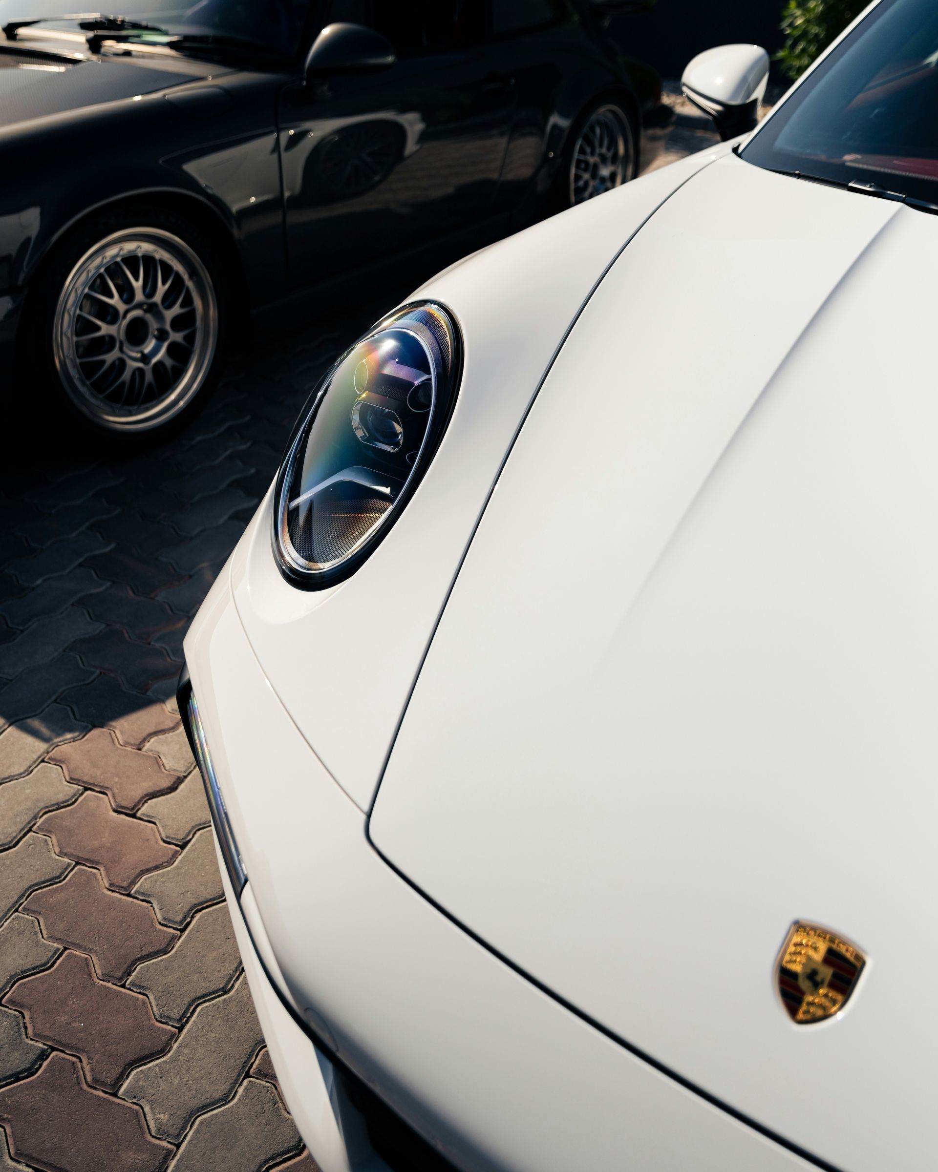 White Porsche sports car front corner with logo, parked beside a black car on brick pavement.
