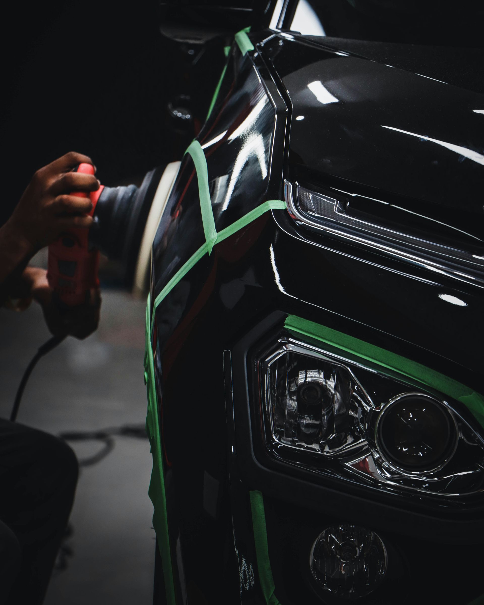 Person polishing a black car’s front fender with green masking tape in a dark workshop