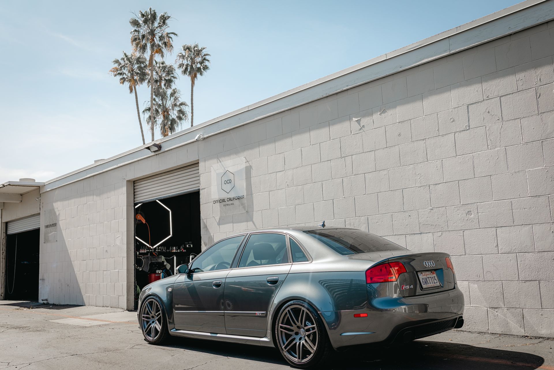 Blue sports coupe parked beside a white cinderblock building with an open garage door and palm trees behind it