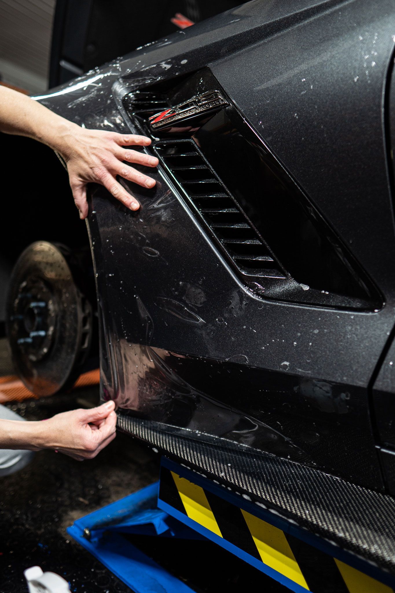 Hands working on a black carbon-fiber car body panel in a garage, near blue jack stands.