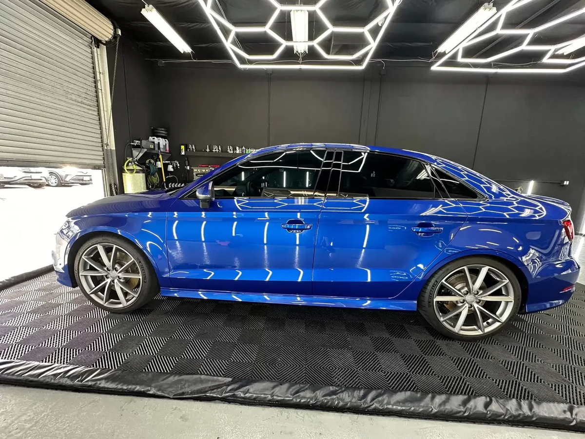 Blue sedan in a modern showroom, viewed from the side on a black patterned floor.