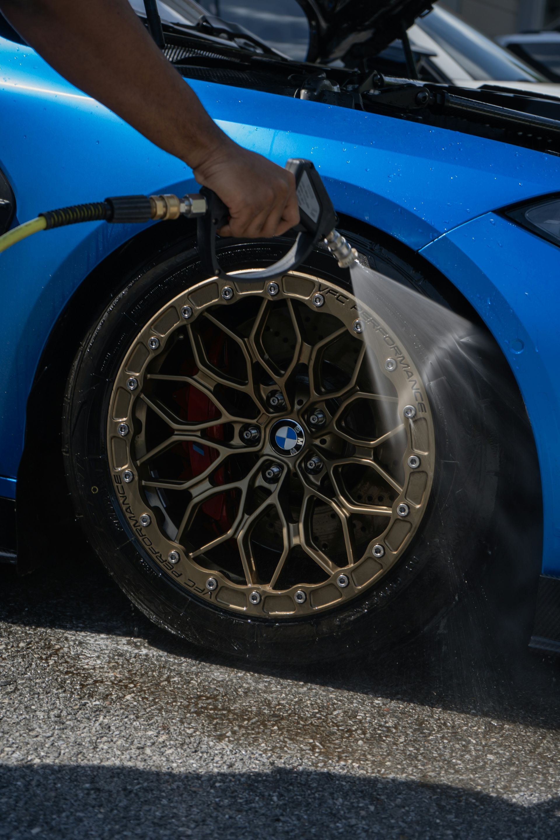 Person sprays water on a blue car’s front wheel with a hose.