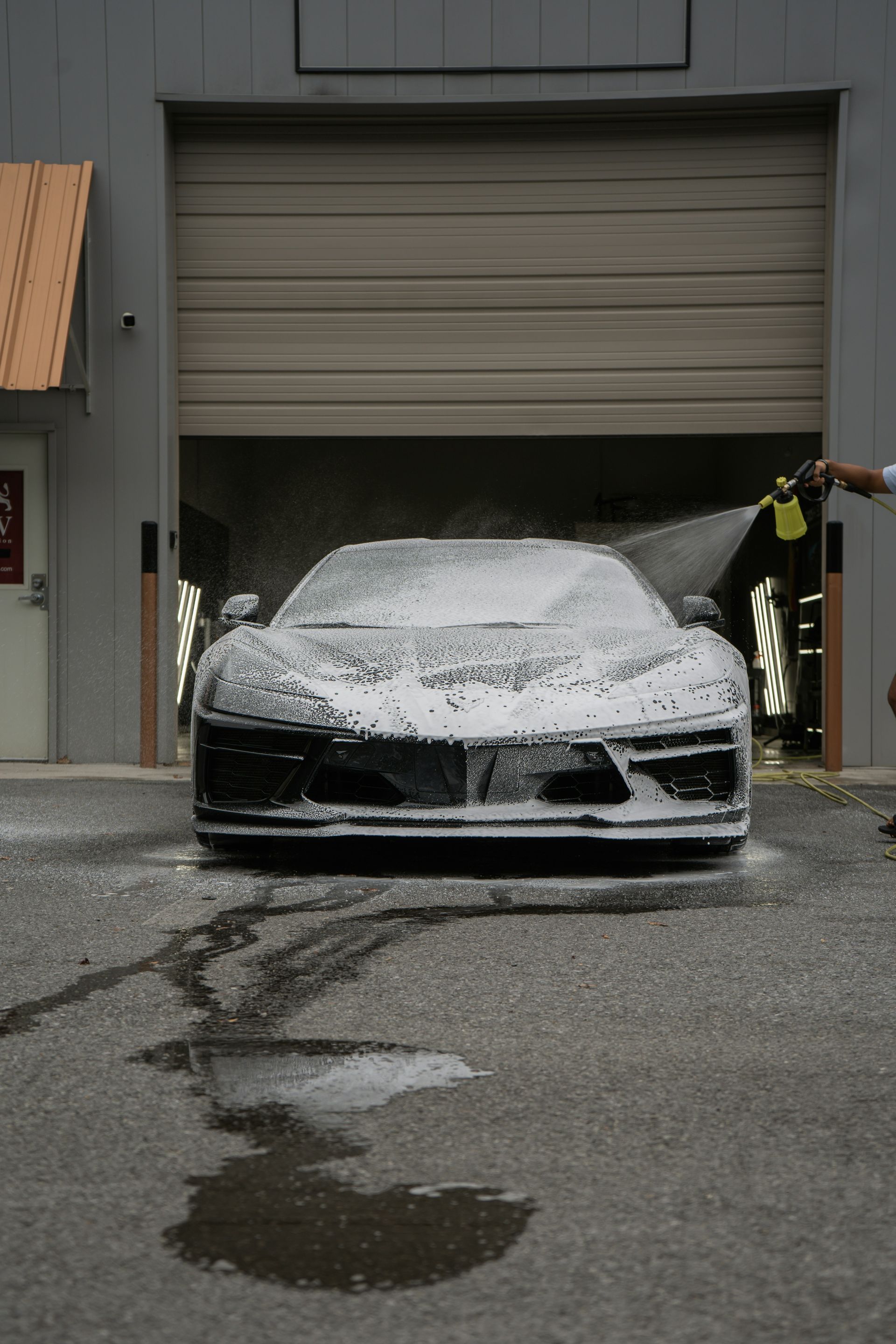Car covered in foam at a garage entrance, with water and suds on the wet pavement.