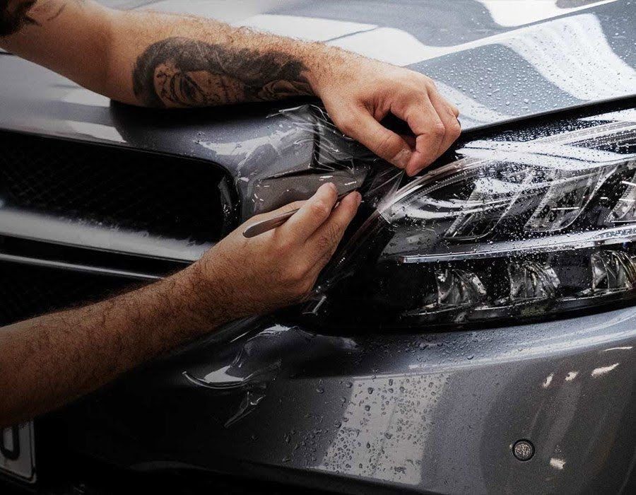 Hands applying a clear protective film to a gray car headlight, with water droplets visible.