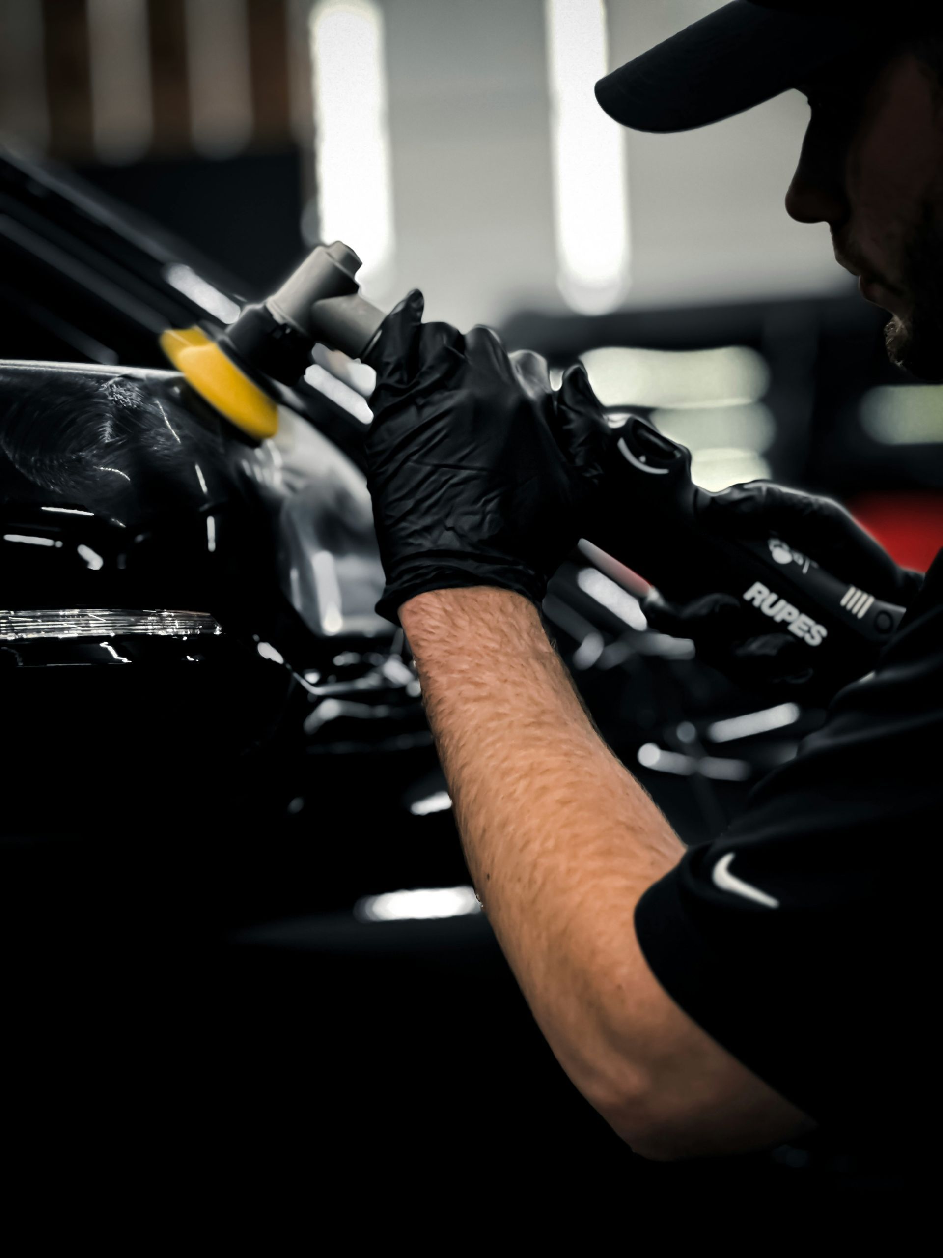 Mechanic in black gloves polishing a car body with a yellow buffing tool in a dark garage