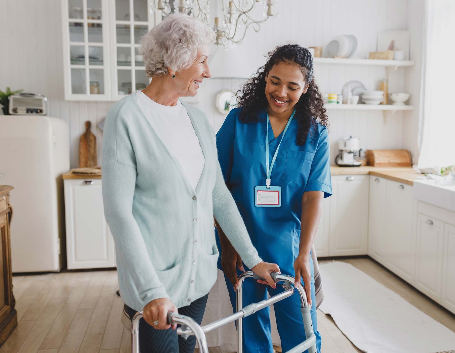Woman assists an older adult using a walker in a kitchen.