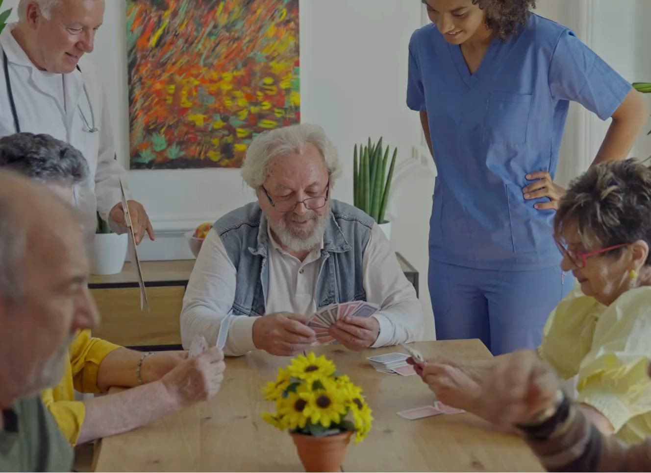 Persone che giocano a carte attorno a un tavolo nella luminosa sala comune di una casa di riposo, con un vaso di fiori gialli al centro.