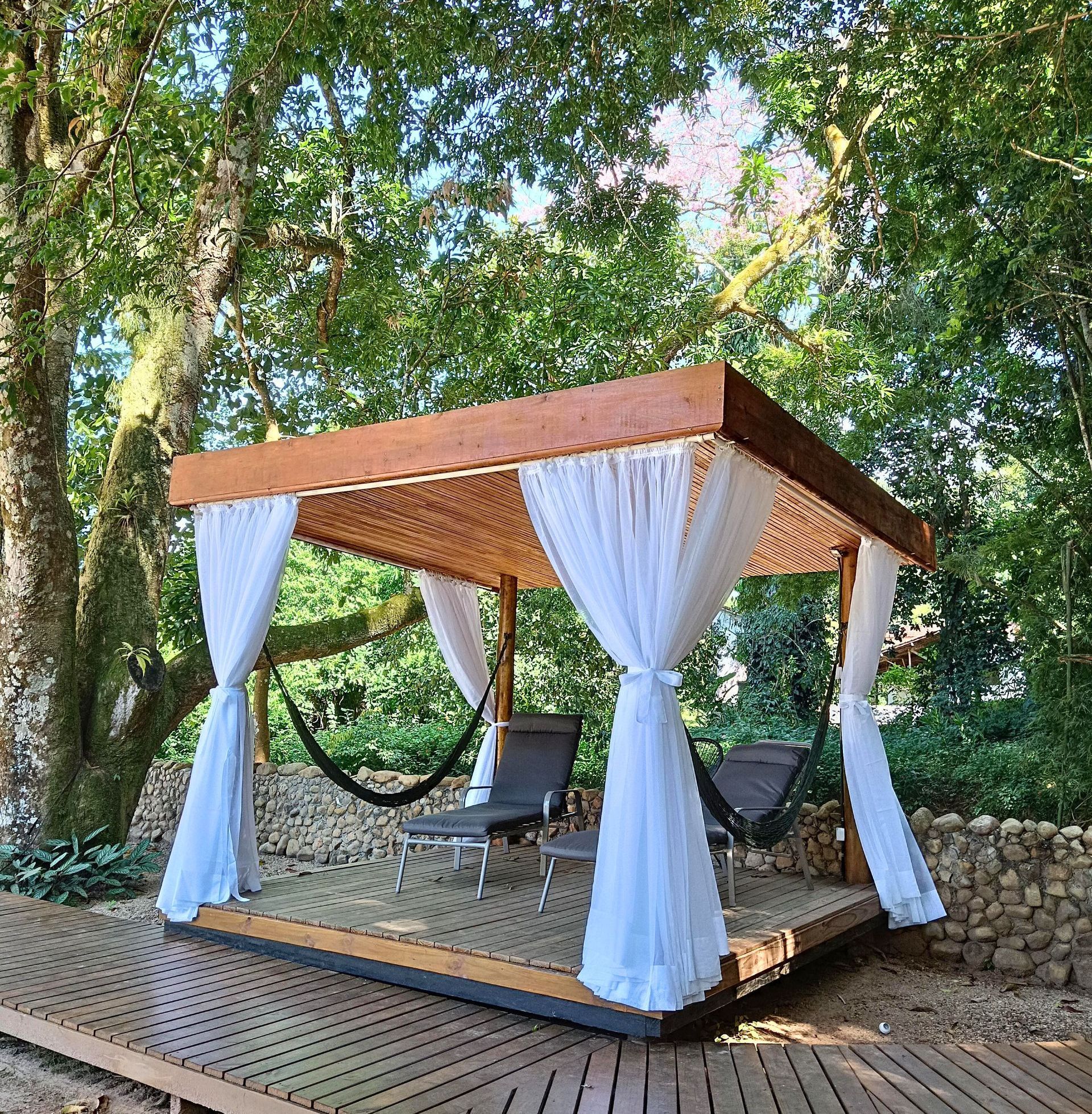 Um gazebo de madeira com redes e cadeiras na floresta.
