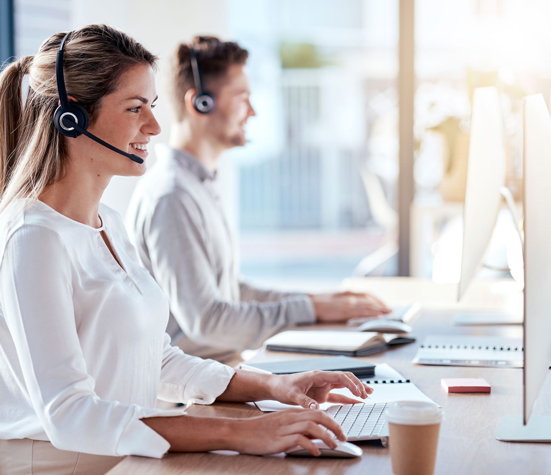 Woman and man with headsets work at computers in a bright office.