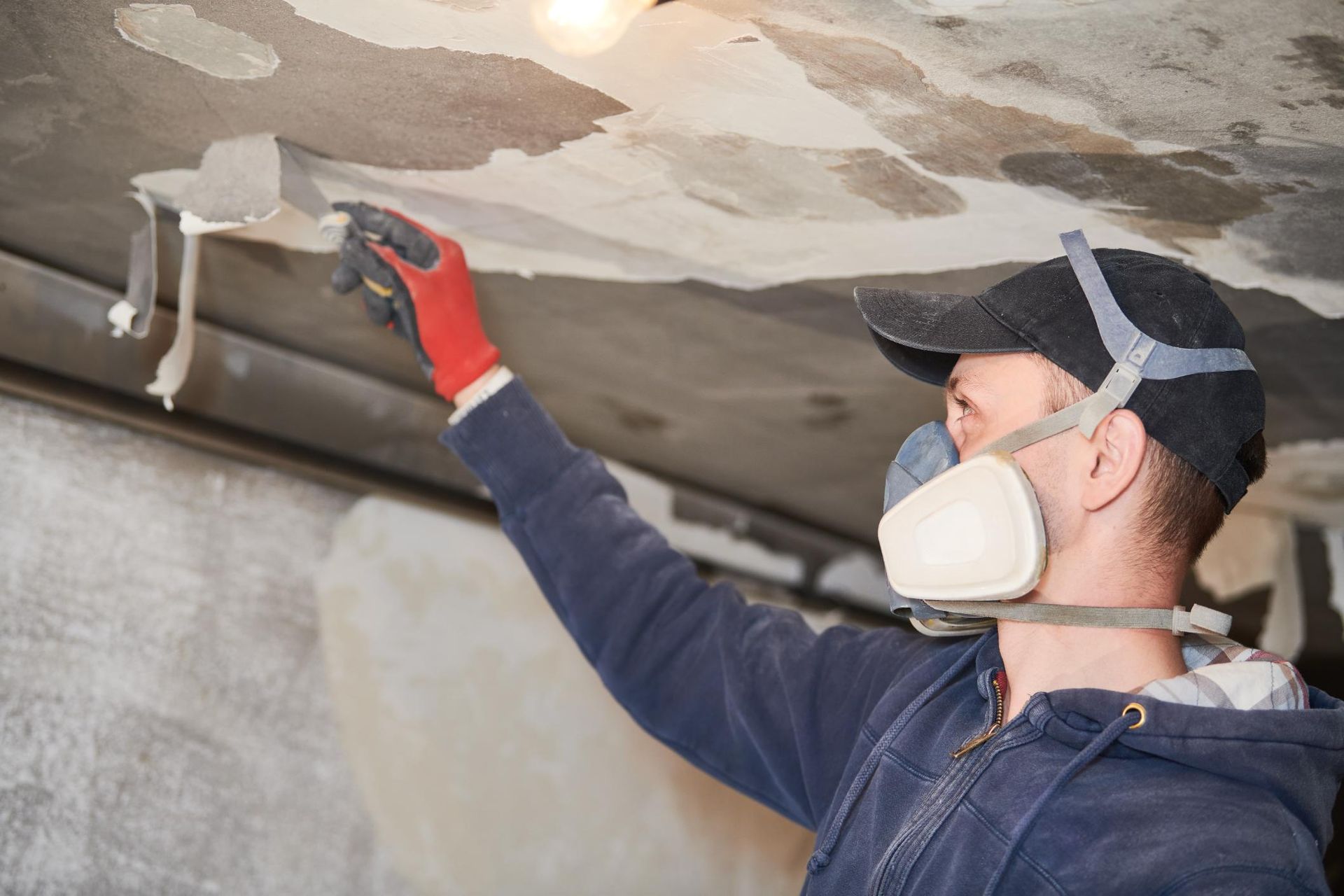 Man scraping peeling paint from ceiling wearing a respirator and gloves.
