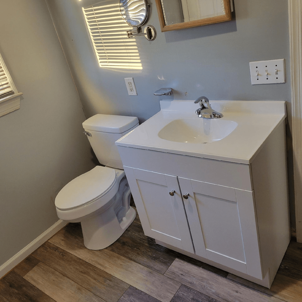 Bathroom with white vanity, toilet, and wood-look flooring; light blue walls, window with blinds.