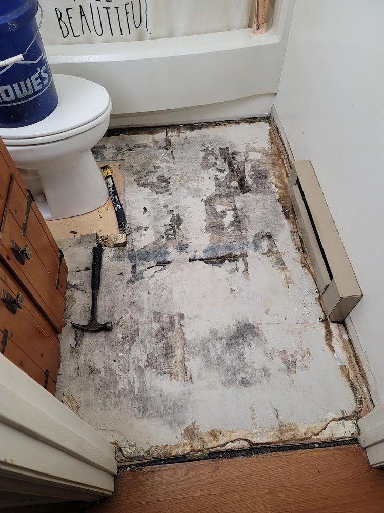 Bathroom with floor damage. Partially removed flooring reveals concrete. Hammer and wood visible.