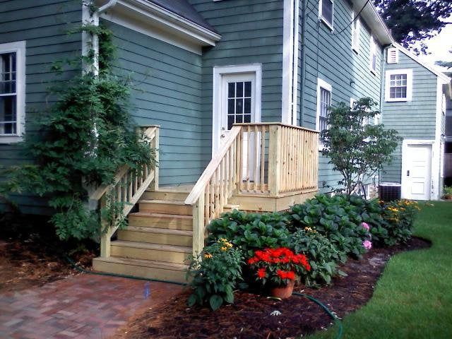 Front steps with wooden railing leading to a green house, bordered by shrubs and red flowers