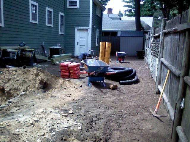 Backyard construction site with dirt trench, wheelbarrow, pipes, and stacked materials along a fence