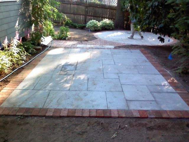 Stone patio with brick border leading to a shaded backyard path and trees