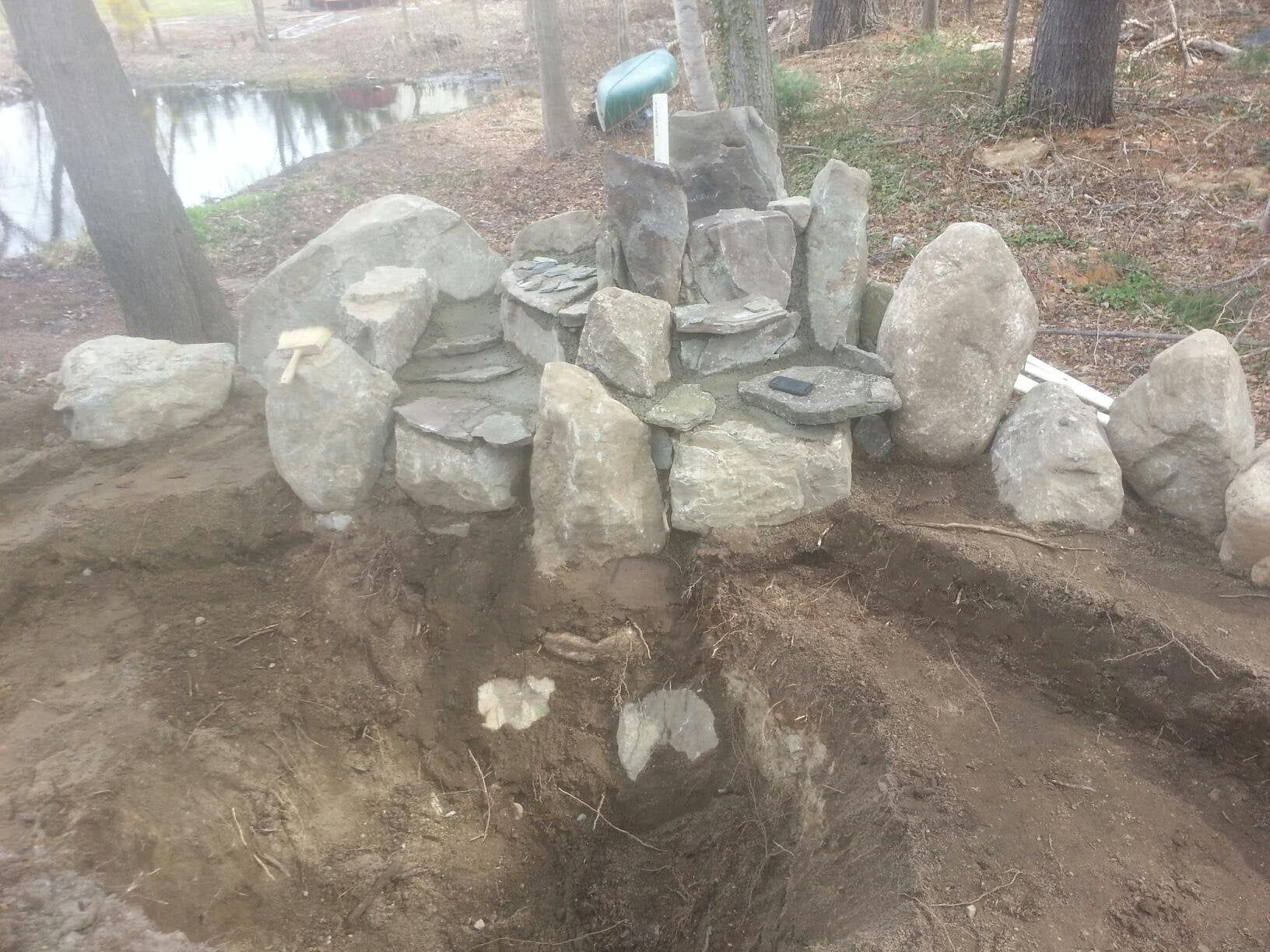 Stone cairn in a wooded area beside a dirt path, with a small blue marker on top.
