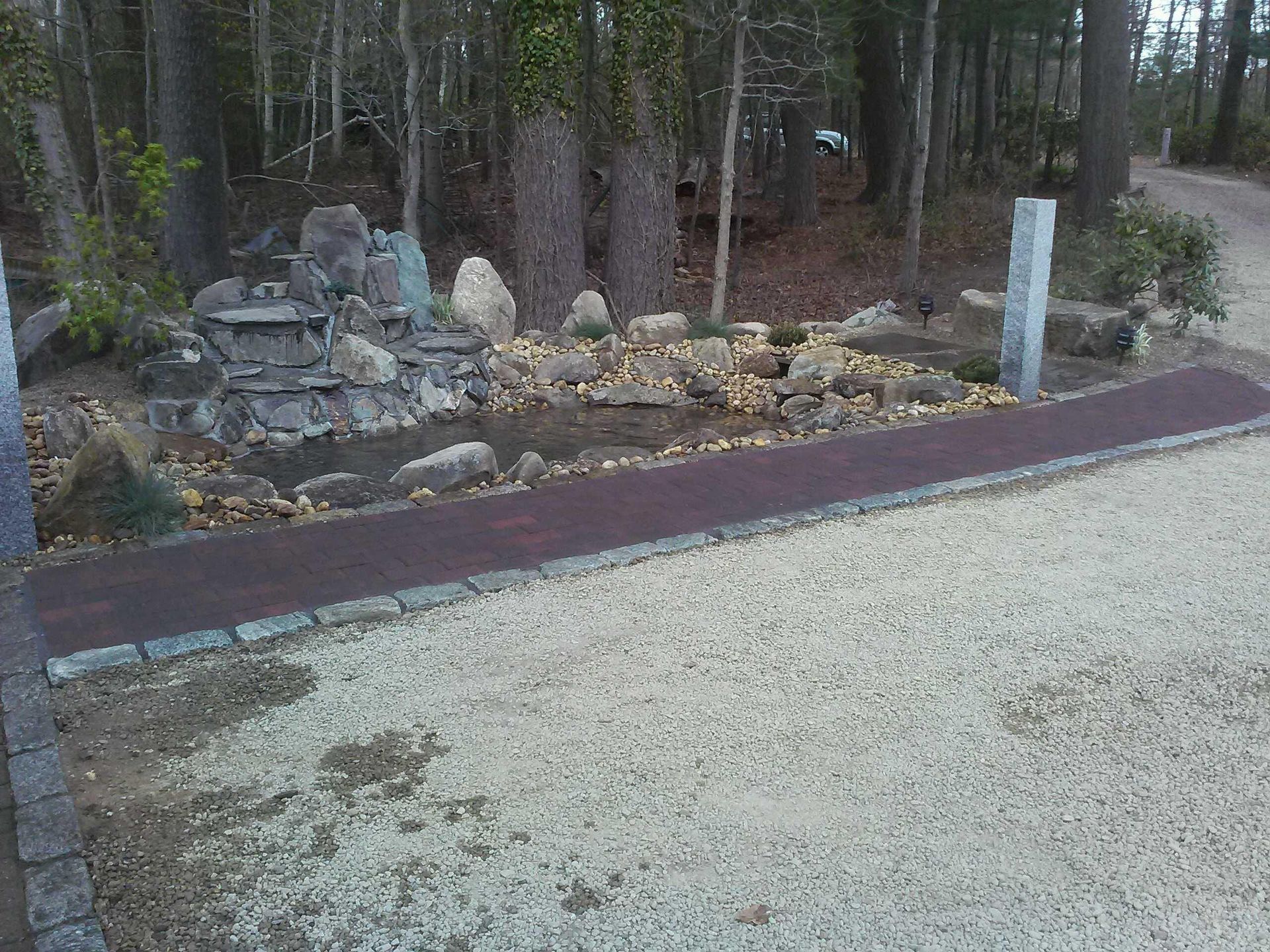 Wooded roadside with a small stone shrine and white markers beside a gravel path
