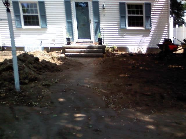 Front yard with a dirt path leading to a house entrance, flanked by piles of soil and a red wheelbarrow.