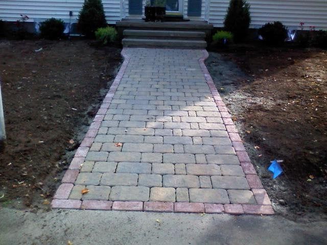 Brick walkway leading to front steps of a house, bordered by bare dirt and small shrubs.