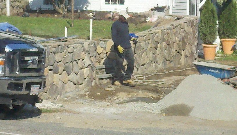 Worker in dark clothes stands by a stone wall at a construction site, with a truck and gravel pile nearby.