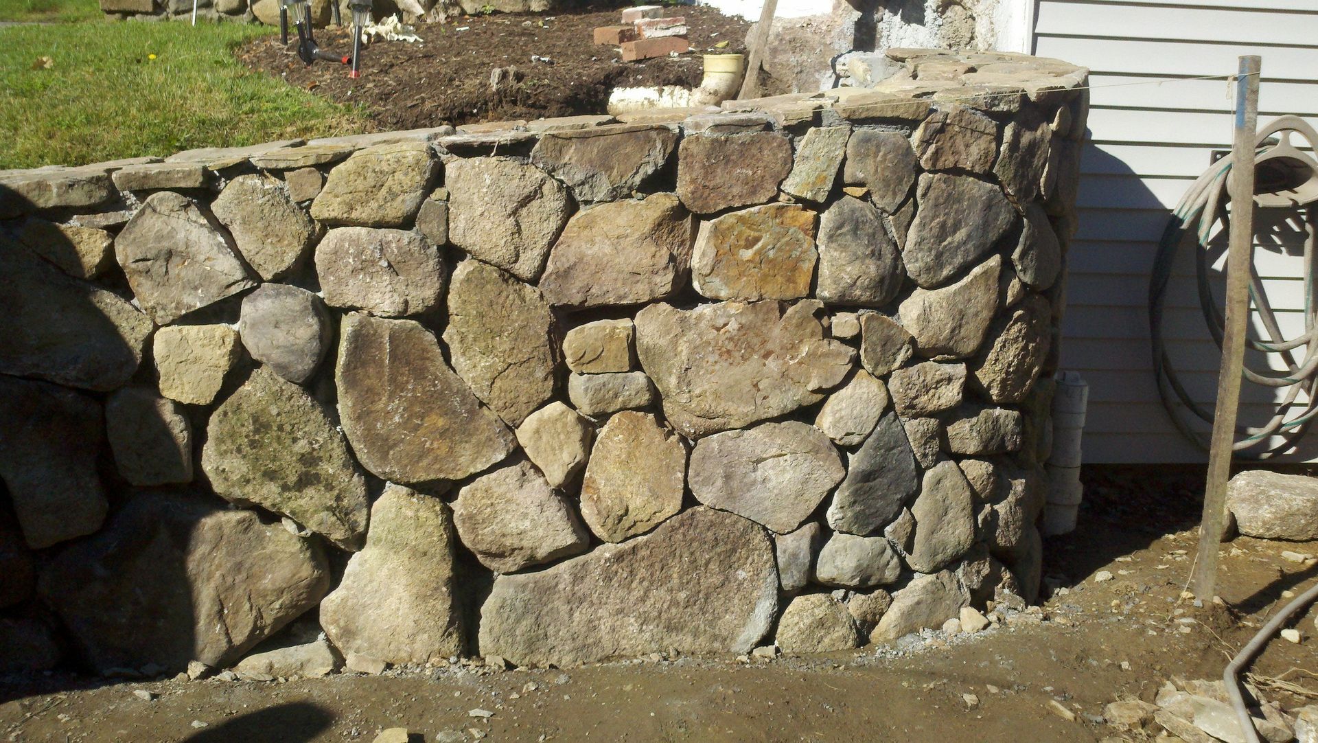 Rough stone retaining wall in a backyard beside a wooden fence and dirt ground