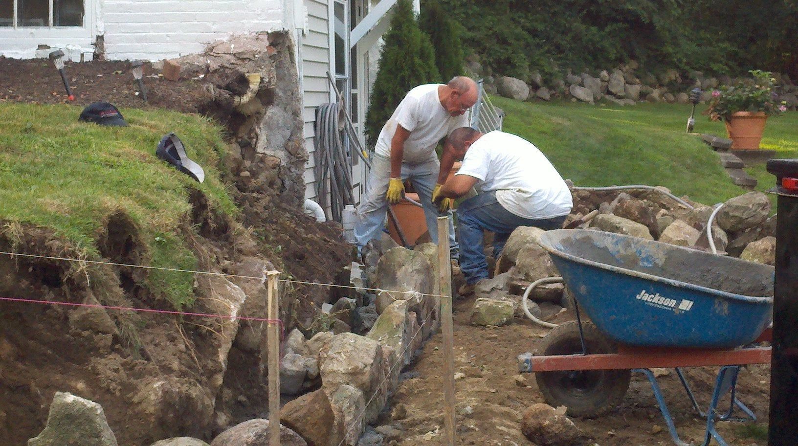 Two workers digging a trench beside a house, with a blue wheelbarrow nearby.