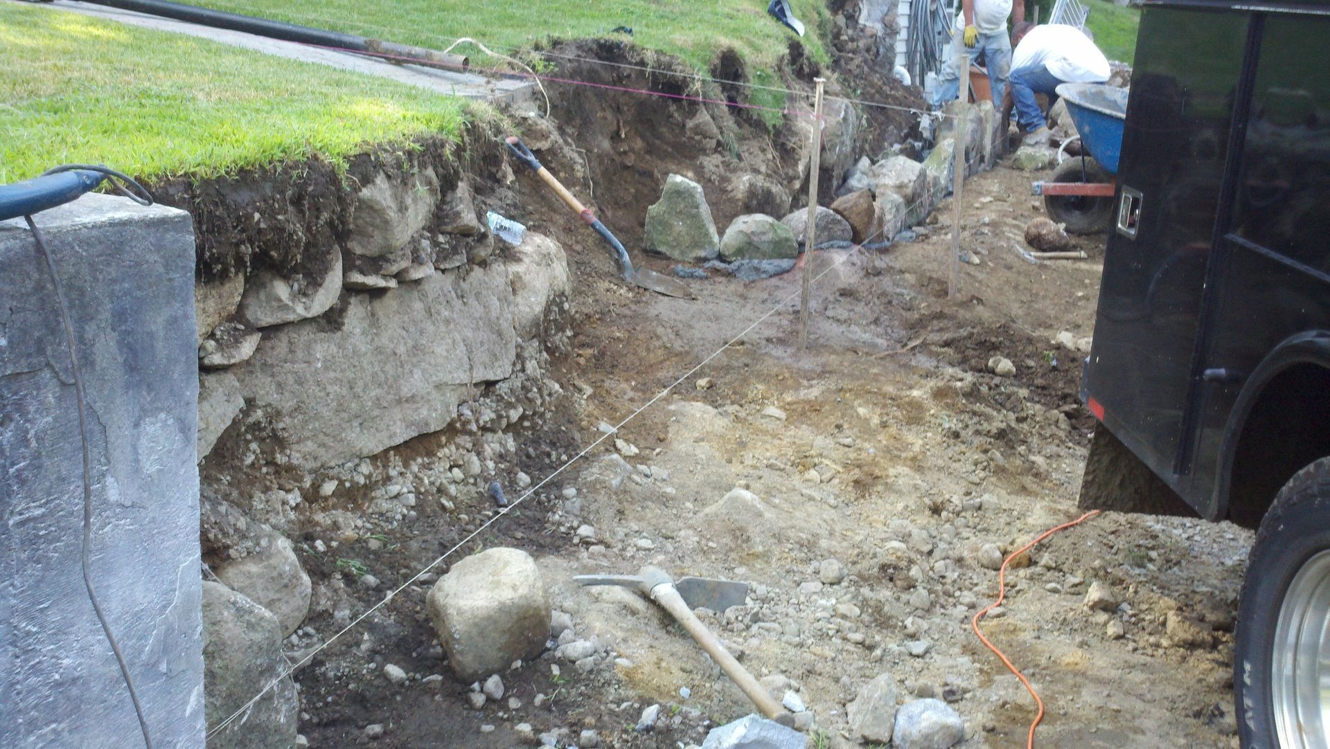 Construction trench beside a stone wall and grassy slope, with workers and a black van nearby.