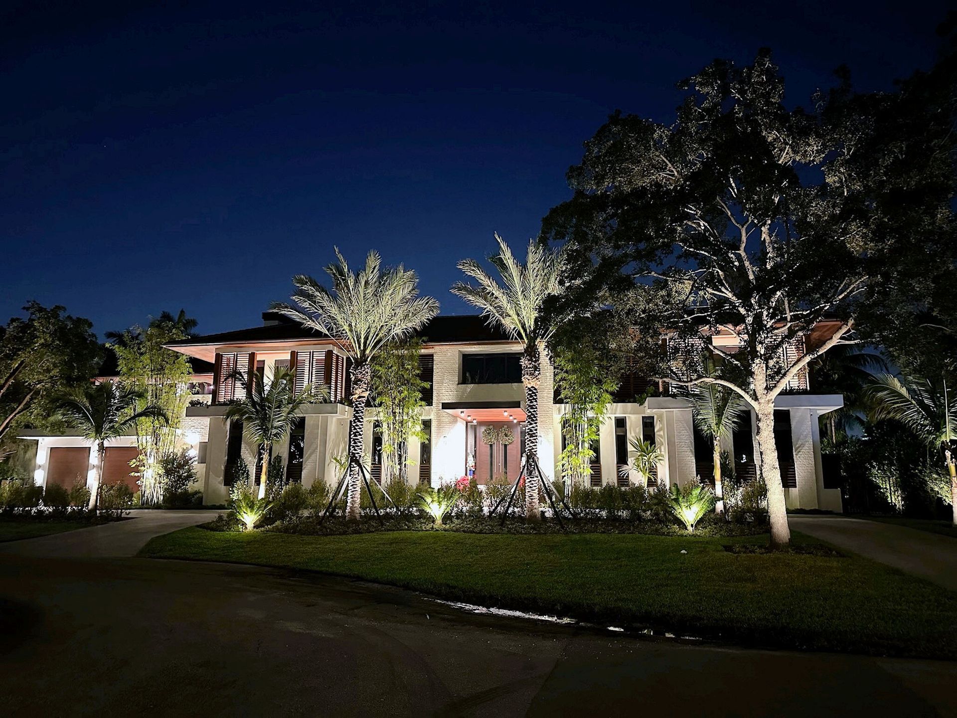 A two-story luxury home at night, illuminated by warm landscape lighting, with illuminated palm trees in the front yard.