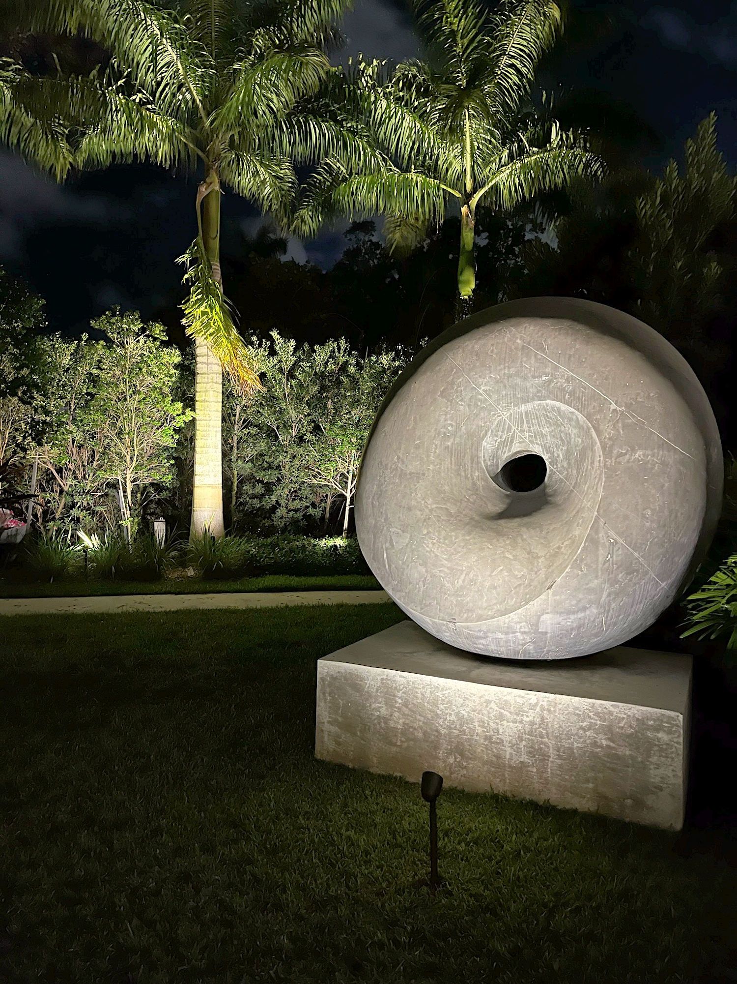 A large, round, stone sculpture with a central hole sits on a square base in a landscaped garden at night.