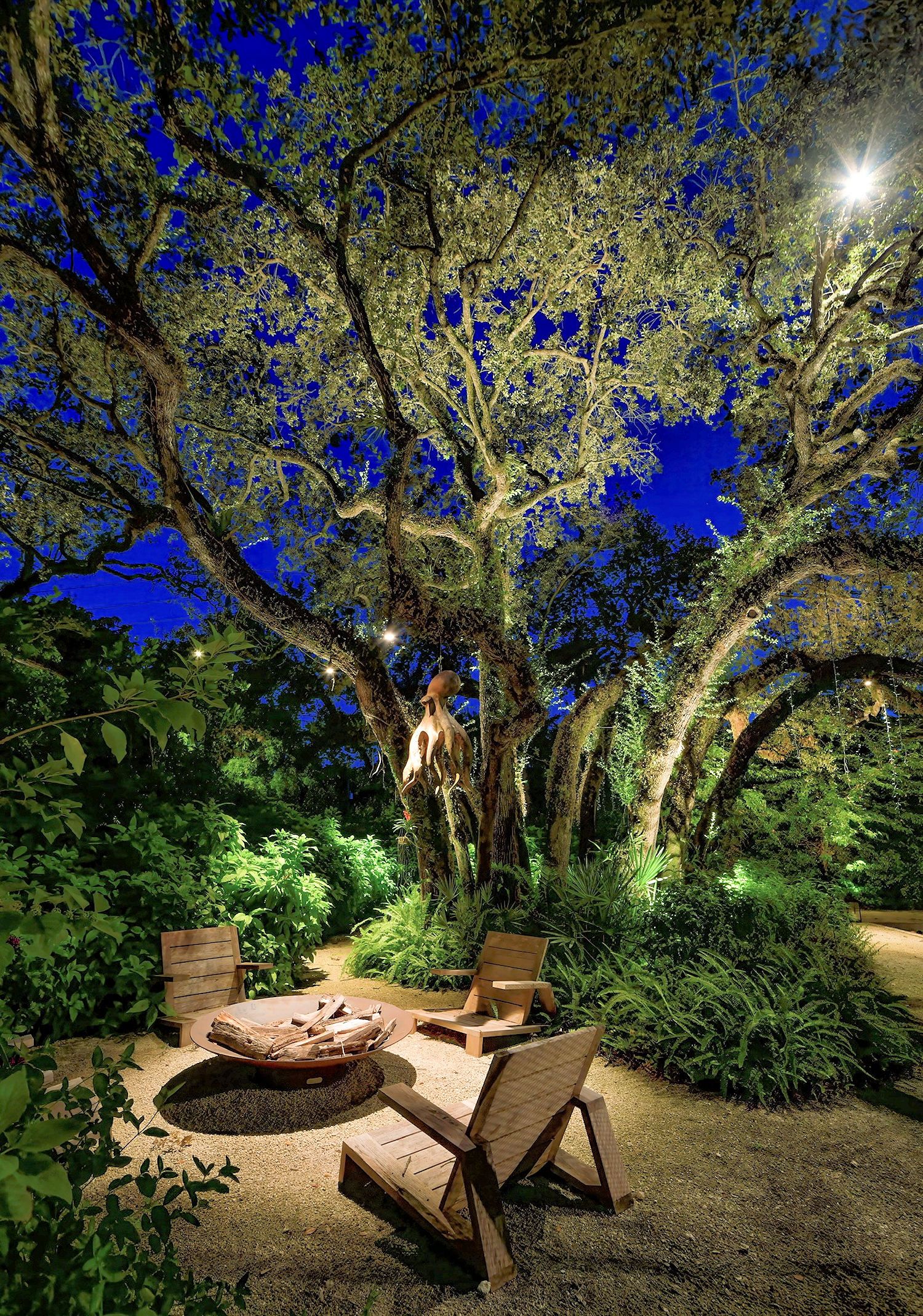 Two wooden lounge chairs sit around a stone fire pit under a large, illuminated tree in a lush garden at night.