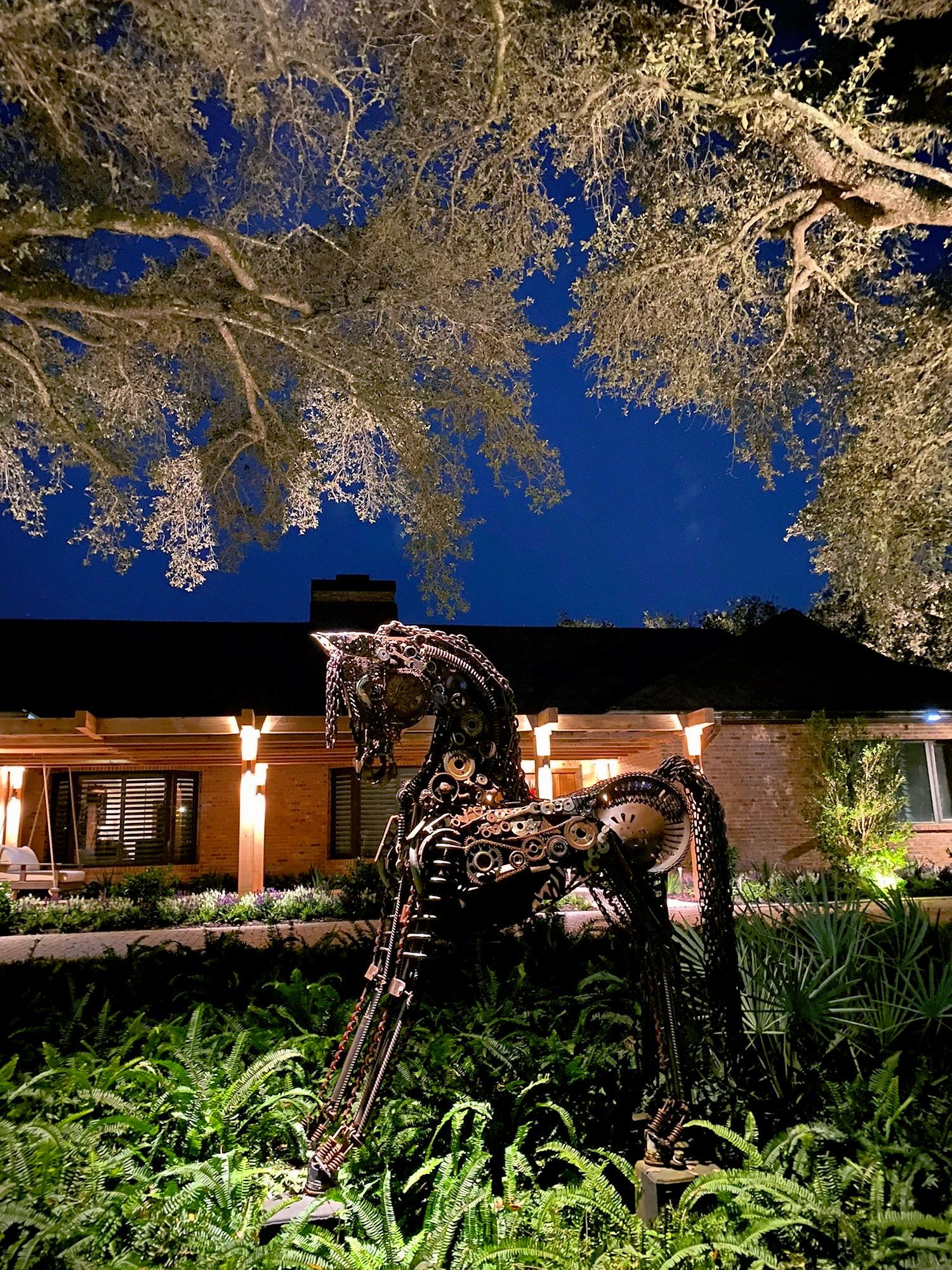 A metal sculpture of a horse stands in a garden at night in front of a house illuminated by warm, glowing lights.