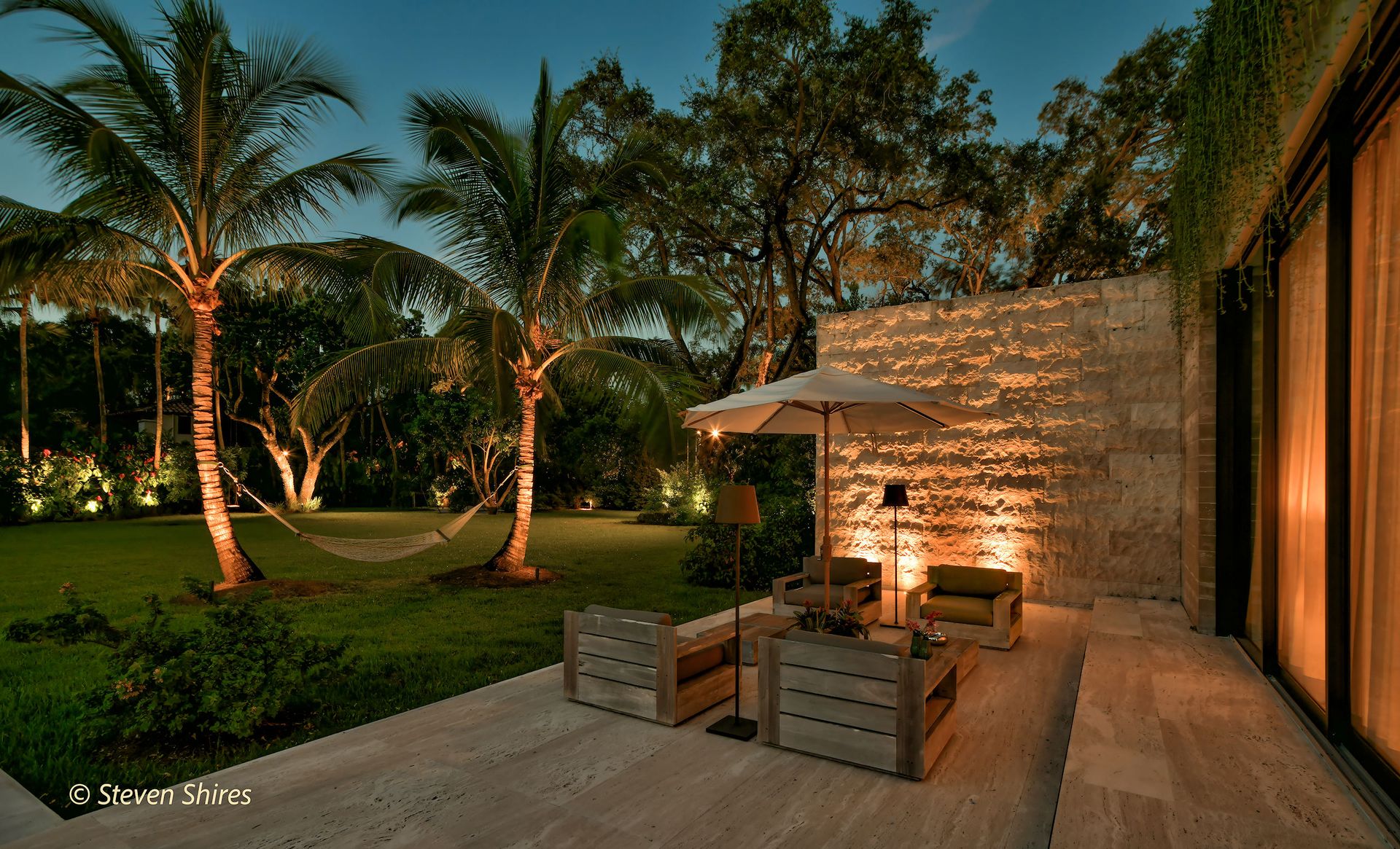A stone patio with outdoor seating and an umbrella at twilight, set against a lush, illuminated garden with palm trees.