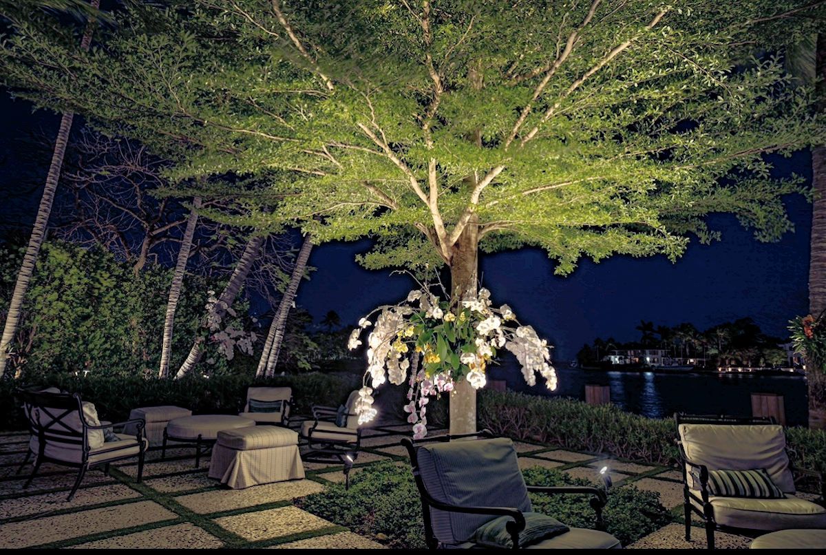 An illuminated patio at night with lounge chairs arranged around a tree decorated with hanging flowers near a waterfront.